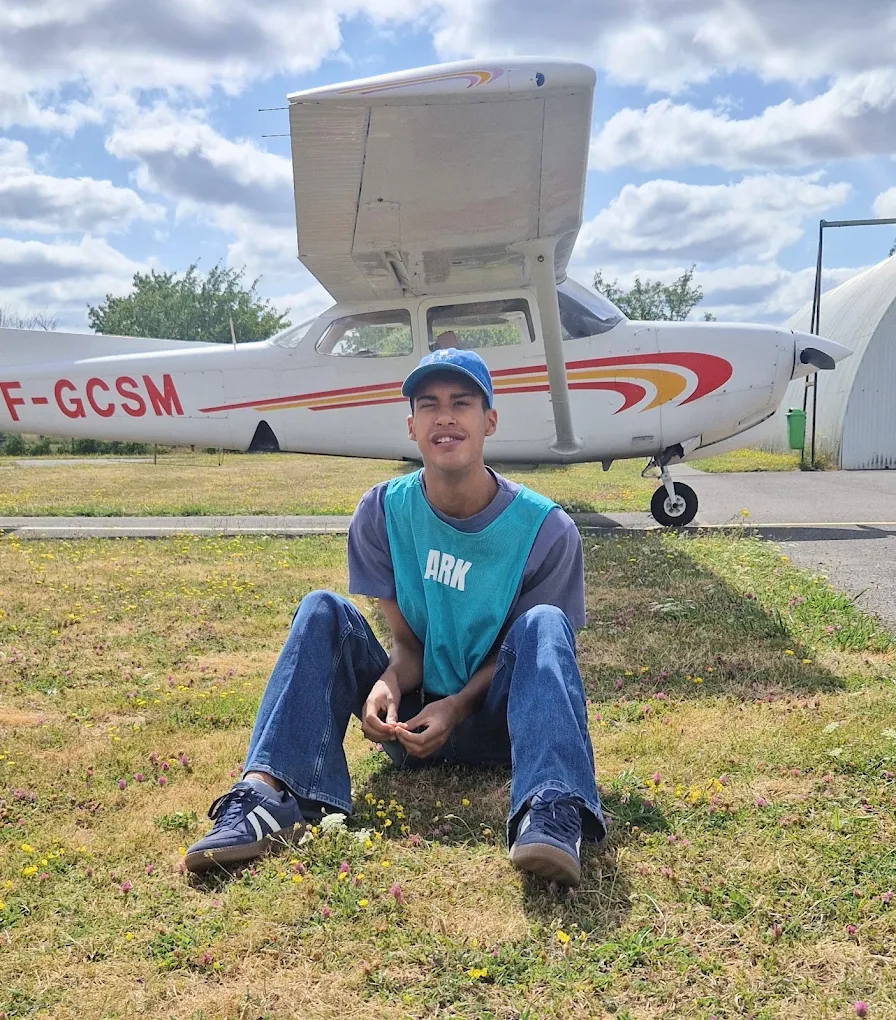 Jeune souriant assis dans l'herbe devant un avion lors d'une sortie découverte